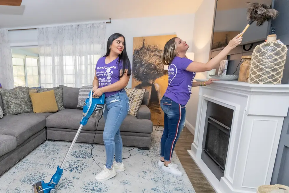 Two CleanFlorida cleaners in purple uniform shirts vacuuming a rug and dusting a white mantel